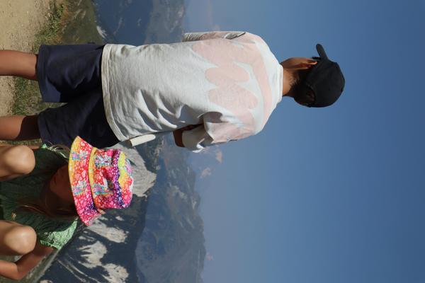 Two individuals stand on a mountain ridge, gazing out at a breathtaking panorama of distant peaks under a clear blue sky